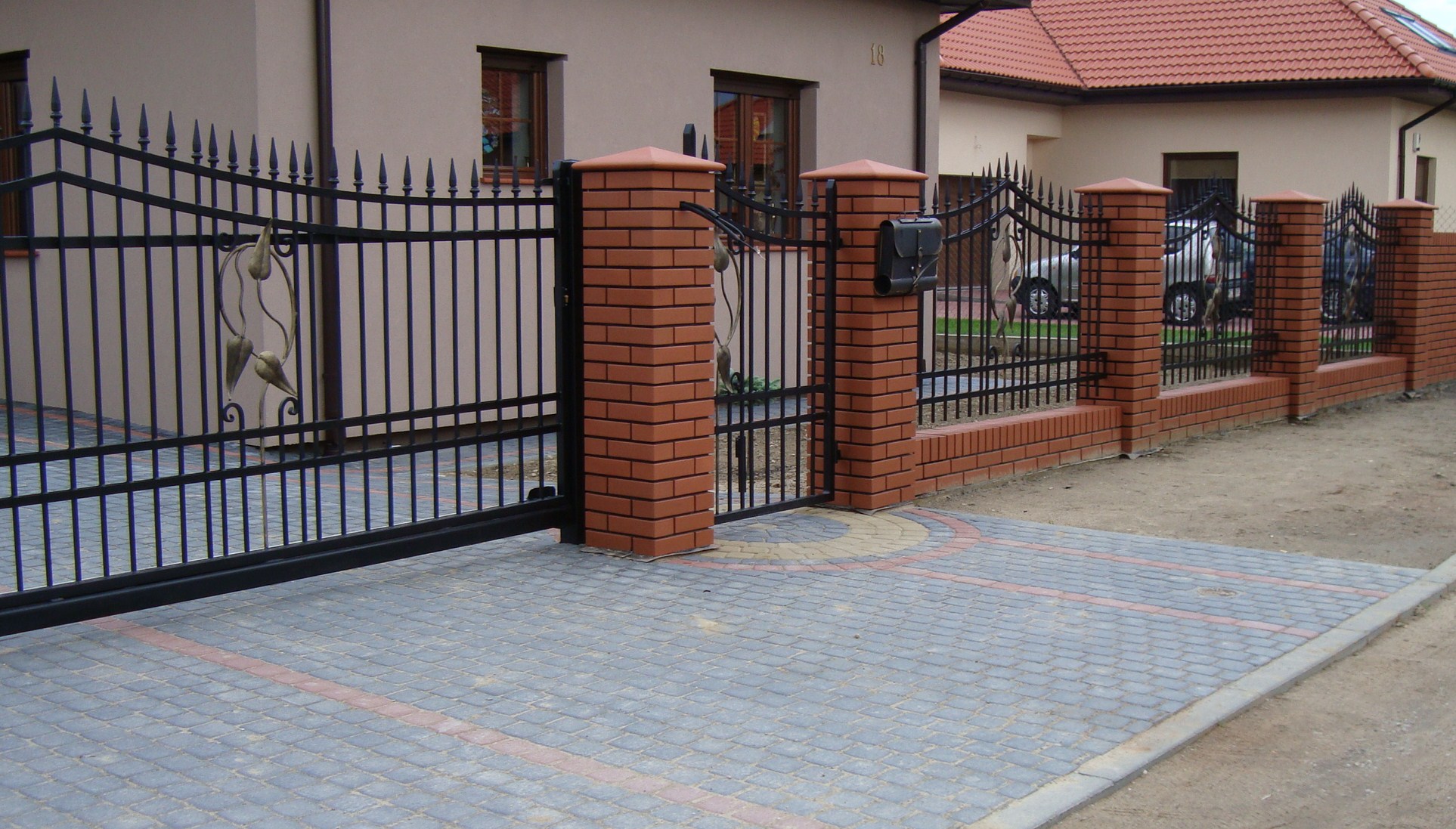 Czarna brama wjazdowa z dekoracyjnymi elementami roślinnymi i furtka z ceglanymi słupkami, widok na posesję z kostki brukowej.